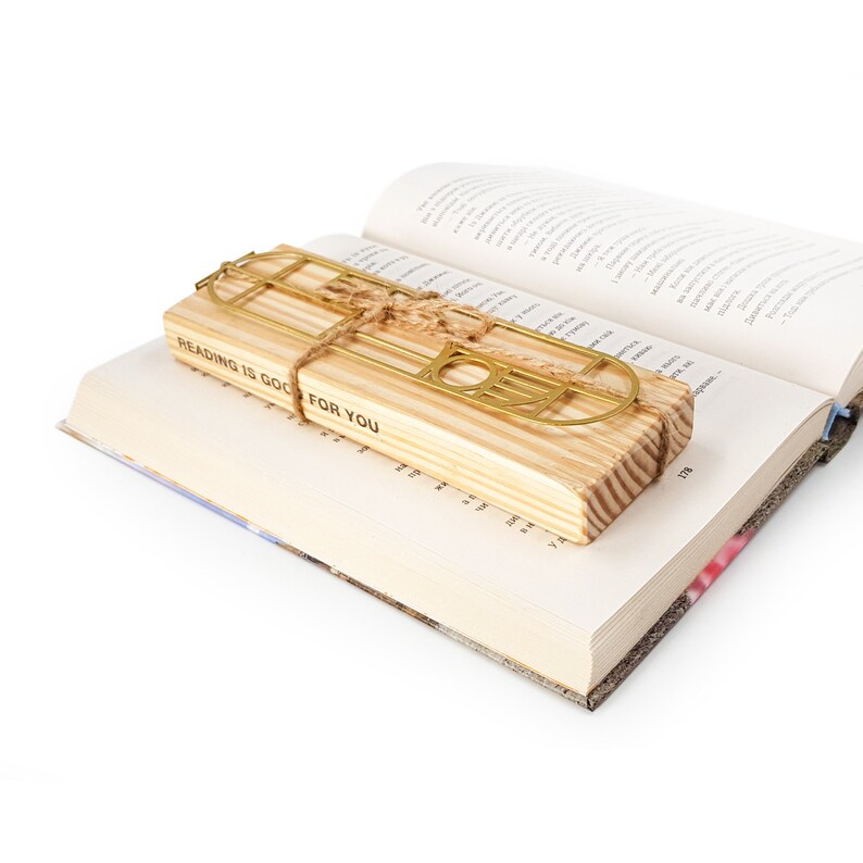 Gold Bauhaus-style metal bookmark tied to a striped wooden block engraved with “READING IS GOOD FOR YOU,” resting on an open book — artistic gift packaging.