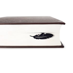 May include: A close-up of a brown hardcover book with a black feather bookmark. The bookmark is placed on the edge of the book.