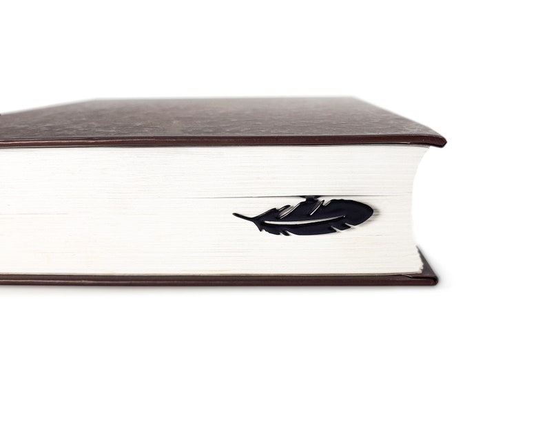 May include: A close-up of a brown hardcover book with a black feather bookmark. The bookmark is placed on the edge of the book.