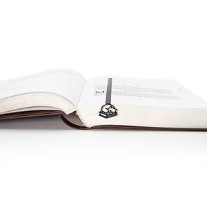 Op de afbeelding: Een zwartmetalen boekenlegger met een kat die op een stapel boeken slaapt. De boekenlegger is in een open boek met witte pagina's en een bruine kaft geplaatst. De afbeelding is tegen een witte achtergrond.