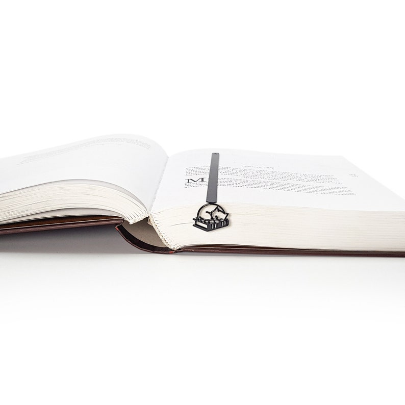 Op de afbeelding: Een zwartmetalen boekenlegger met een kat die op een stapel boeken slaapt. De boekenlegger is in een open boek met witte pagina's en een bruine kaft geplaatst. De afbeelding is tegen een witte achtergrond.
