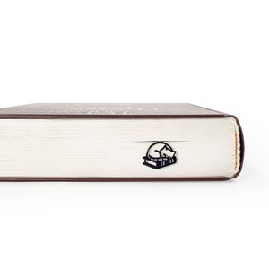 Op de afbeelding: Zwart metalen boekenlegger met een slapende kat op twee boeken. De boekenlegger is in een dik boek gestoken met witte pagina's en een bruine kaft. De afbeelding is tegen een witte achtergrond.