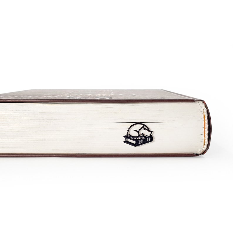 Op de afbeelding: Zwart metalen boekenlegger met een slapende kat op twee boeken. De boekenlegger is in een dik boek gestoken met witte pagina's en een bruine kaft. De afbeelding is tegen een witte achtergrond.