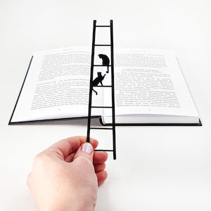 Black metal cat bookmark with two cats climbing a ladder, held by hand inside an open book, minimalist playful design.