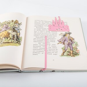 A pink castle bookmark with a pink ribbon. The bookmark is on top of an open book with illustrations of people riding horses.