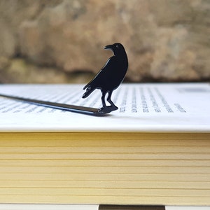 Black metal raven bookmark standing upright on the pages of a closed book, with a stone wall in the background. A sleek gothic gift for lovers of mystery, poetry, and dark literature.