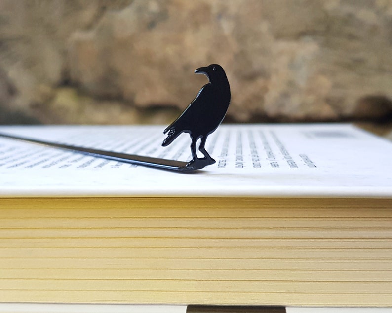 Black metal raven bookmark standing upright on the pages of a closed book, with a stone wall in the background. A sleek gothic gift for lovers of mystery, poetry, and dark literature.