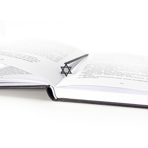 Minimalist black metal Star of David bookmark placed in the spine of an open book, clean white background and soft light for modern aesthetic.