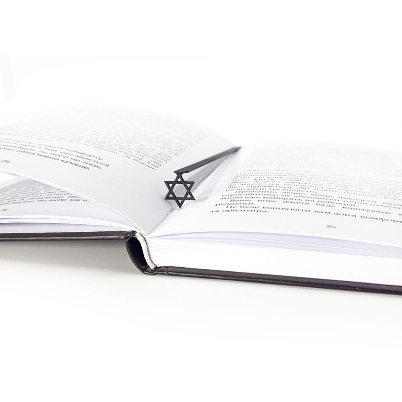 Minimalist black metal Star of David bookmark placed in the spine of an open book, clean white background and soft light for modern aesthetic.