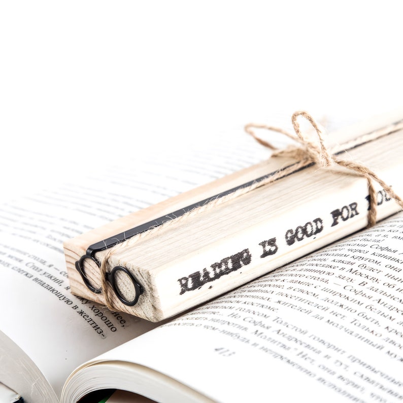Architect-inspired metal page saver shaped like round glasses, wrapped on a wooden block stamped with ‘Reading is good for you,’ resting inside an open book. Minimalist Bauhaus-style gift for modern readers.