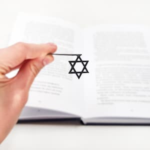Hand holding a black metal Star of David bookmark over an open book, minimal and modern design, perfect for book lovers and symbolic gifts.