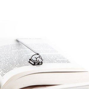 Op de afbeelding: Zwart metalen boekenlegger met een slapende kat ontwerp, geplaatst in een open boek. Het silhouet van de kat is zichtbaar tussen de pagina's. Het paginanummer van het boek, 195, is ook zichtbaar.