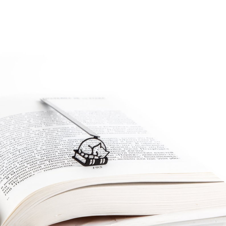 Op de afbeelding: Zwart metalen boekenlegger met een slapende kat ontwerp, geplaatst in een open boek. Het silhouet van de kat is zichtbaar tussen de pagina's. Het paginanummer van het boek, 195, is ook zichtbaar.