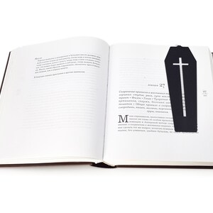Black metal coffin-shaped bookmark with a cross cutout placed inside an open book, lying flat across the right-hand page.