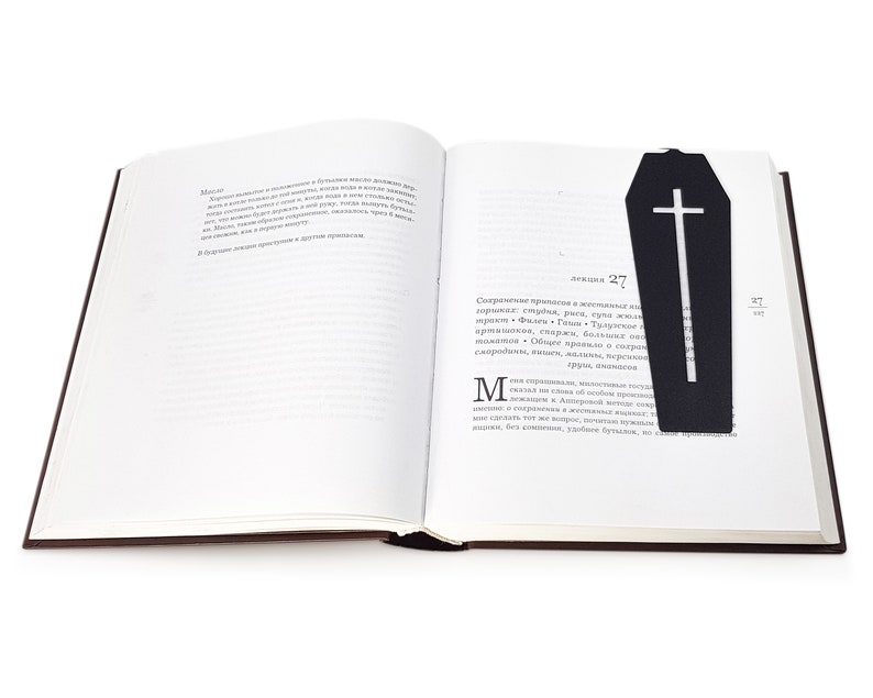 Black metal coffin-shaped bookmark with a cross cutout placed inside an open book, lying flat across the right-hand page.