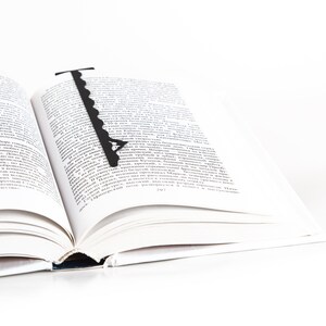 May include: A black bookmark shaped like a key with a decorative design is placed in an open book. The book is open to a page with text in a foreign language.
