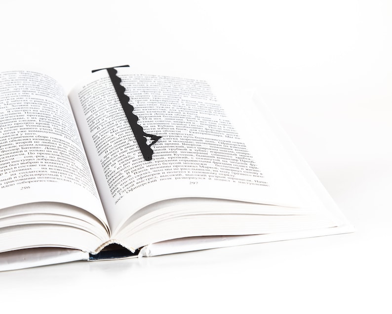 May include: A black bookmark shaped like a key with a decorative design is placed in an open book. The book is open to a page with text in a foreign language.
