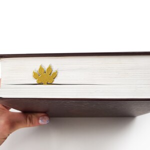 May include: A closed book with a brown cover and a gold paw print bookmark. The book is being held by a hand.