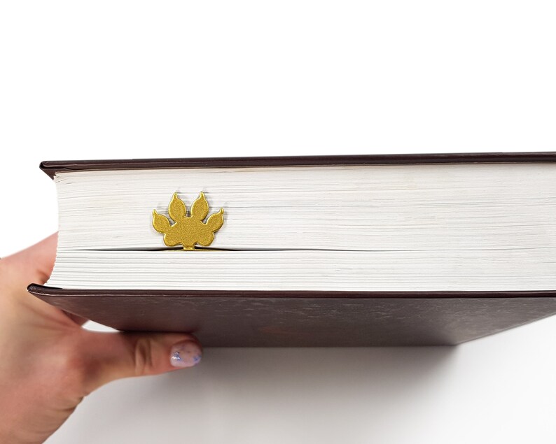 May include: A closed book with a brown cover and a gold paw print bookmark. The book is being held by a hand.