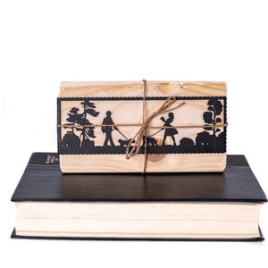 May include: A wooden box with a black silhouette of a couple walking with a dog, tied with twine. The box is sitting on top of a black and cream book.
