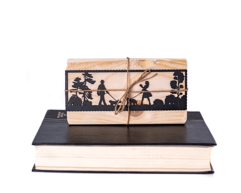 May include: A wooden box with a black silhouette of a couple walking with a dog, tied with twine. The box is sitting on top of a black and cream book.