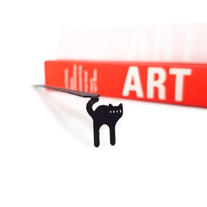 Minimalist-style black metal bookmark Amazing Kitty. A flat metal stick is sitting between the pages of the book. The amazing can is seen on the side of the book even when its closed.