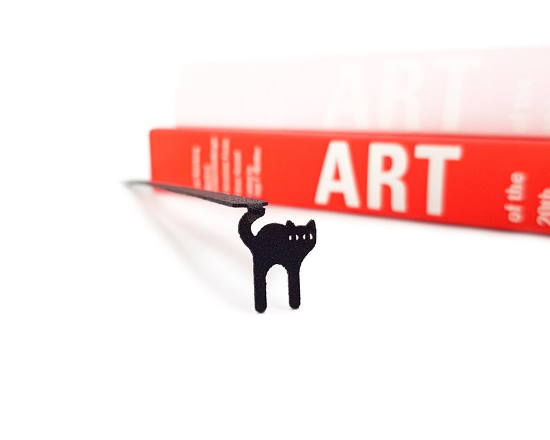Minimalist-style black metal bookmark Amazing Kitty. A flat metal stick is sitting between the pages of the book. The amazing can is seen on the side of the book even when its closed.