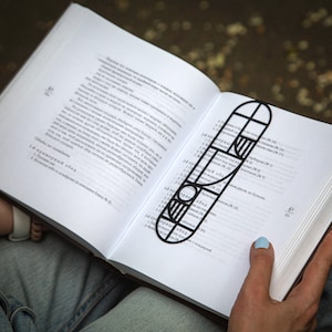 Person reading a book with a black metal Bauhaus-style bookmark resting on the open pages; geometric design in minimalist modern style. The bookmark is black metal with a geometric design. The bookmark is shaped like a long rectangle with rounded edges. The design features a combination of circles, squares, and stripes. The bookmark is placed in a book with white pages and black text. Designed and made by Coolbookmark in Ukraine.