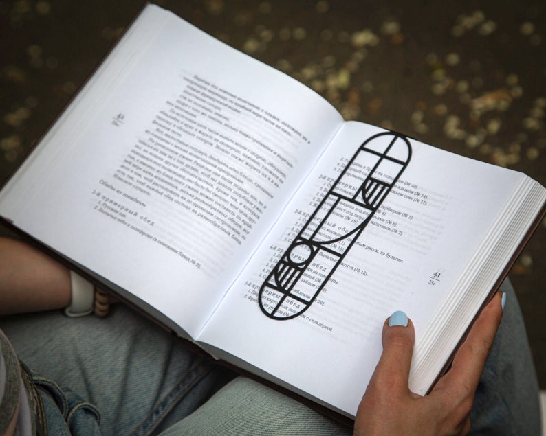 Person reading a book with a black metal Bauhaus-style bookmark resting on the open pages; geometric design in minimalist modern style. The bookmark is black metal with a geometric design. The bookmark is shaped like a long rectangle with rounded edges. The design features a combination of circles, squares, and stripes. The bookmark is placed in a book with white pages and black text. Designed and made by Coolbookmark in Ukraine.