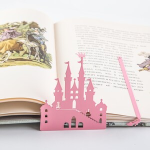 A pink metal castle bookmark with a decorative design. The bookmark is placed on top of an open book with a white background.