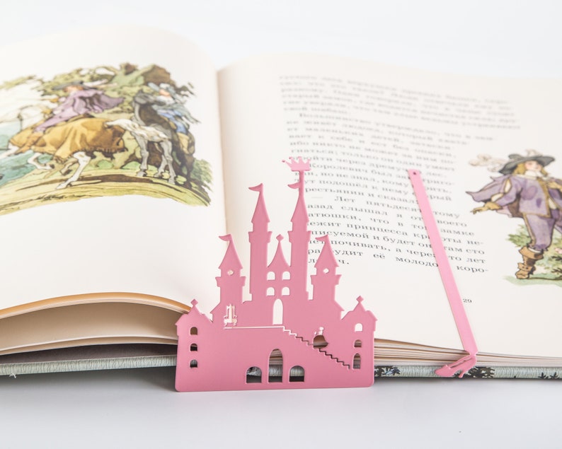 A pink metal castle bookmark with a decorative design. The bookmark is placed on top of an open book with a white background.
