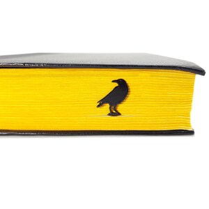 Black metal page saver shaped like a raven, tucked into the yellow-edged pages of a closed book. A moody, literary gift for Poe fans and readers.