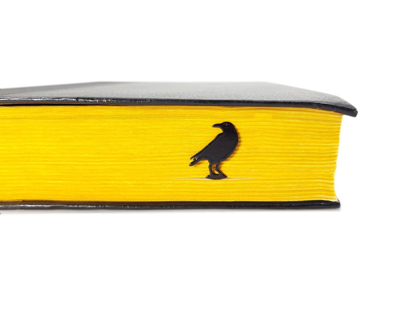 Black metal page saver shaped like a raven, tucked into the yellow-edged pages of a closed book. A moody, literary gift for Poe fans and readers.