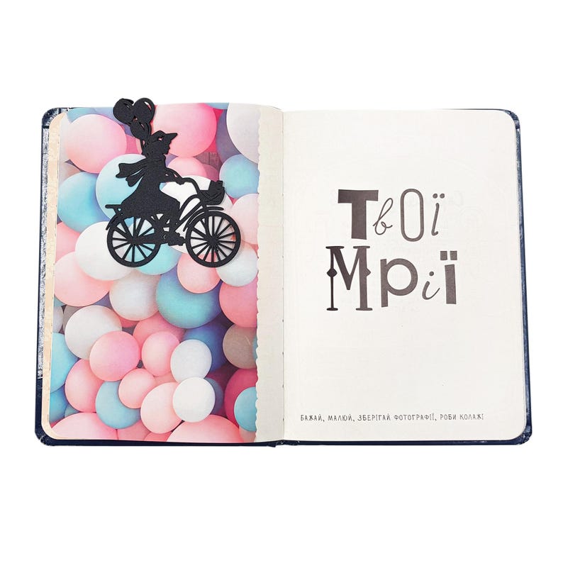 Black metal bike bookmark shaped as a girl riding a bicycle with balloons, placed inside an open book against colorful balloon-patterned endpapers.