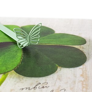 May include: A green metal butterfly bookmark with a four-leaf clover design. The bookmark is on a white background with the word "mbiu" written in cursive.