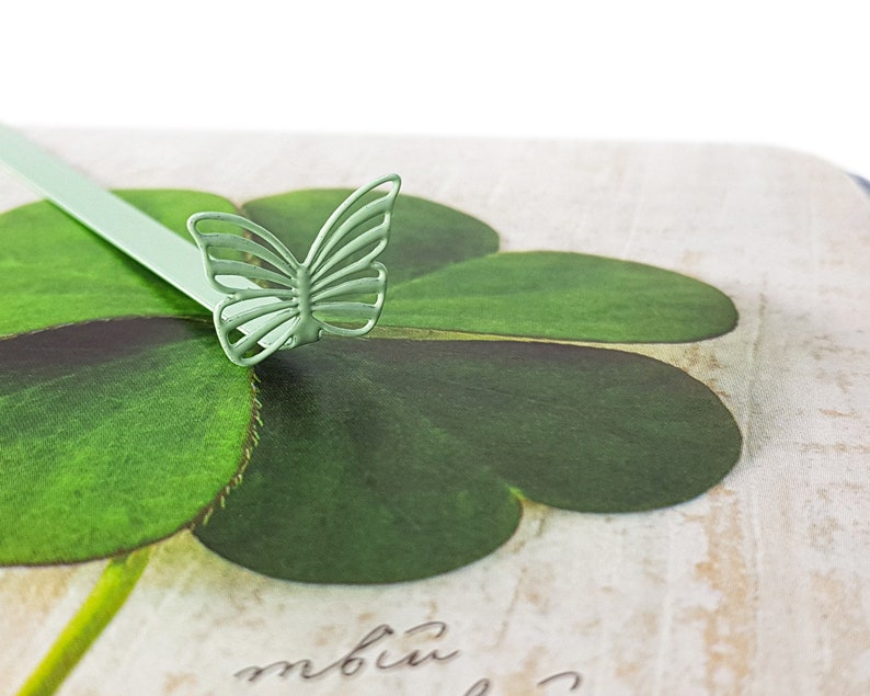 May include: A green metal butterfly bookmark with a four-leaf clover design. The bookmark is on a white background with the word "mbiu" written in cursive.