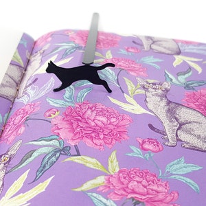 Black metal running cat bookmark placed inside a book with purple pages decorated with illustrated cats and pink peony flowers.
