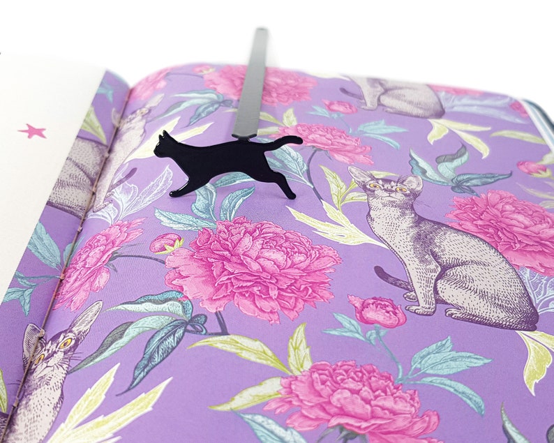 Black metal running cat bookmark placed inside a book with purple pages decorated with illustrated cats and pink peony flowers.