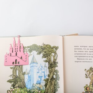 A pink metal castle shaped bookmark on top of an open book with illustrations of a castle and trees.