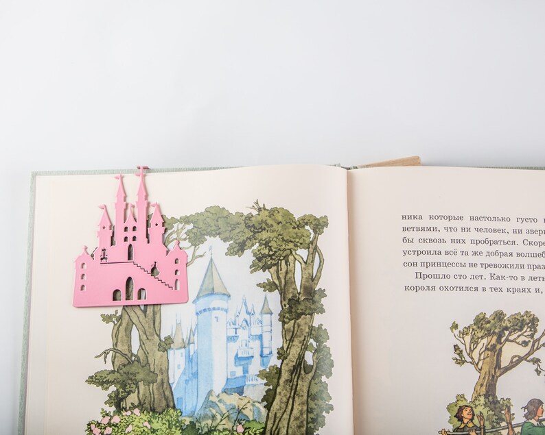 A pink metal castle shaped bookmark on top of an open book with illustrations of a castle and trees.