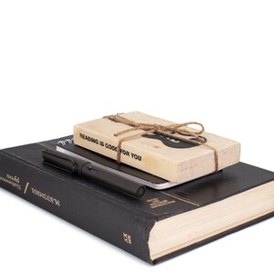 Electric guitar-shaped black metal bookmark tied to a wooden block with the phrase Reading is good for you, shown with a pen and closed hardcover book. A ready-to-gift set for music-loving readers.