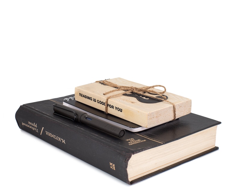 Electric guitar-shaped black metal bookmark tied to a wooden block with the phrase Reading is good for you, shown with a pen and closed hardcover book. A ready-to-gift set for music-loving readers.