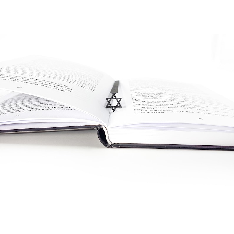 Black Star of David metal bookmark centered in the spine of an open book with white pages and black cover, modern and meaningful reading accessory.