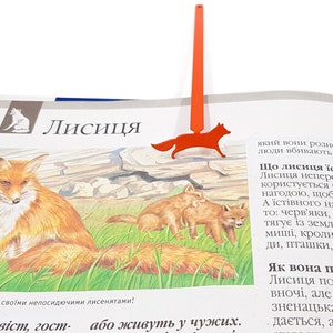 May include: A red fox bookmark with a thin, long, red metal rod. The bookmark is shaped like a fox with a long, bushy tail. The bookmark is on top of a page of a book about foxes. The text on the page reads "Лисиця" which is Ukrainian for "Fox".