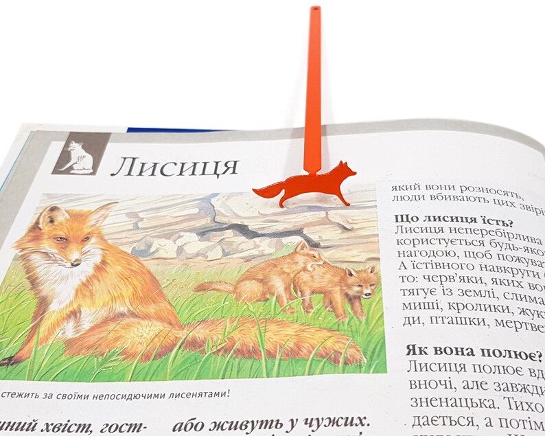 May include: A red fox bookmark with a thin, long, red metal rod. The bookmark is shaped like a fox with a long, bushy tail. The bookmark is on top of a page of a book about foxes. The text on the page reads "Лисиця" which is Ukrainian for "Fox".