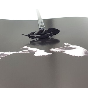 May include: A black metal feather-shaped stamp with a black handle. The stamp is on a black surface with three white silhouettes of birds.