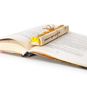 May include: A yellow and brown wooden bookmark with the text "читаю, когда хочу" in black lettering. The bookmark is tied with twine and is placed on an open book.