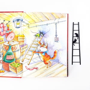 Metal cat bookmark shaped like a ladder with two cats climbing, placed beside an open illustrated book with a colorful cat scene.