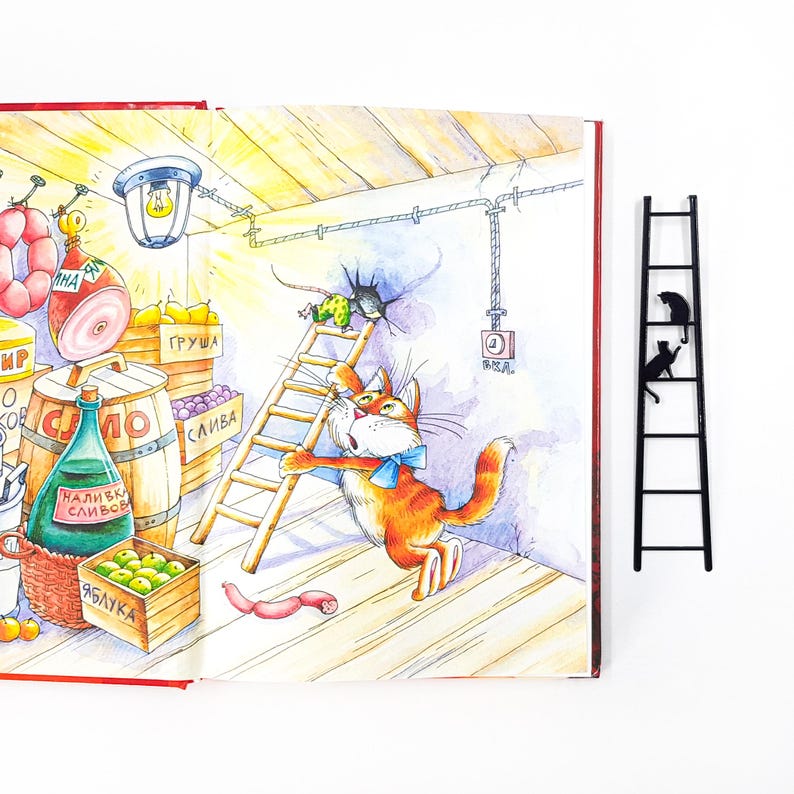 Metal cat bookmark shaped like a ladder with two cats climbing, placed beside an open illustrated book with a colorful cat scene.