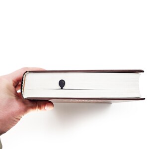 Black metal bike bookmark peeking from the pages of a closed book, showing the balloon detail while the book is held in one hand against a white background.